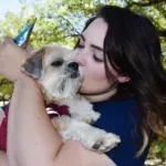 Woman in blue shirt kisses a small, tan dog outdoors under trees.