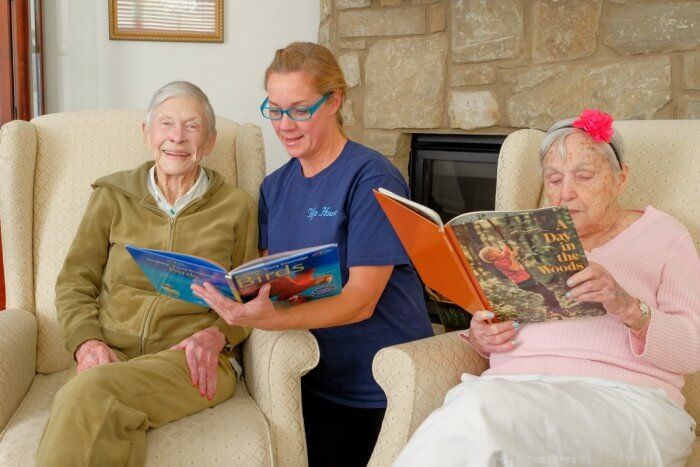 Woman reads aloud to two seniors in armchairs, reading picture books. Fireplace in background.