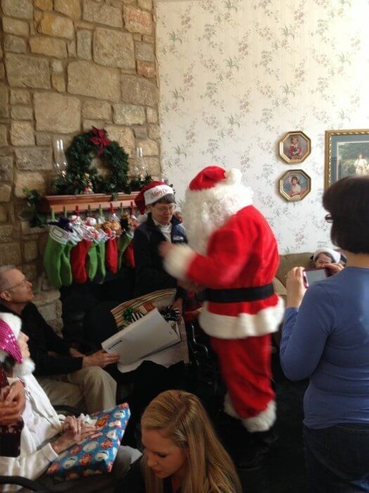 Santa Claus with gifts in a living room, giving to seated people. Decorated fireplace in background.