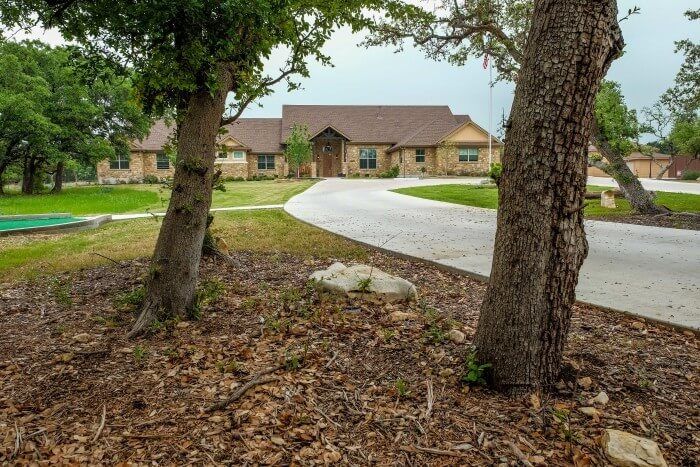 Long, single-story brick home with a winding driveway, framed by trees, on a grassy lot.