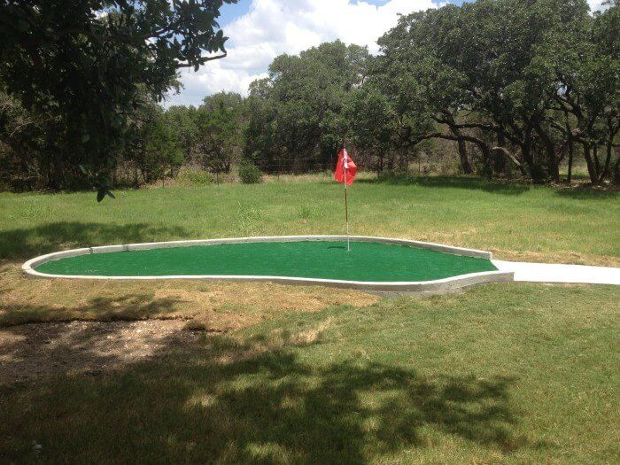 A small green putting green with a red flag, surrounded by grass and trees.