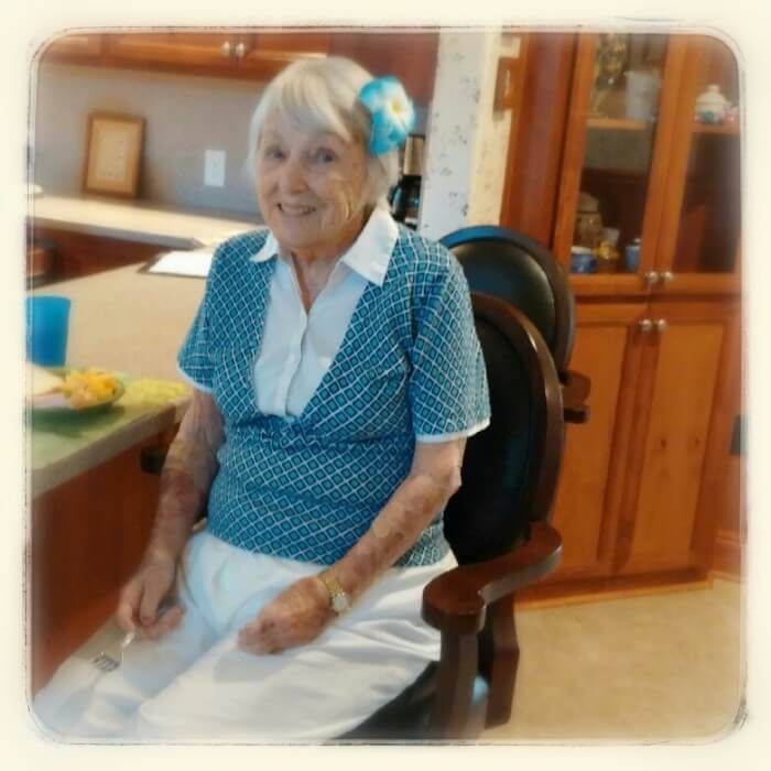 Elderly woman smiling, wearing a blue floral hair clip, sitting indoors near a kitchen counter.