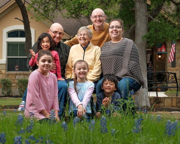 Family portrait: smiling people of various ages pose in front of a house, surrounded by bluebonnets.