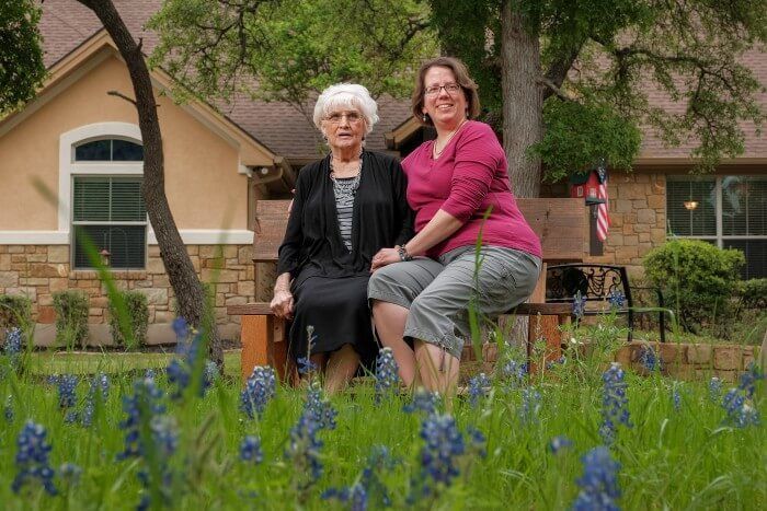 Two women seated in a field of bluebonnets in front of a house. One is smiling, the other is neutral.