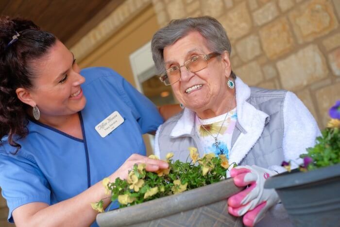 Caregiver and senior woman tending flowers in a pot, outdoors. Woman smiles, wearing gloves; caregiver assists.