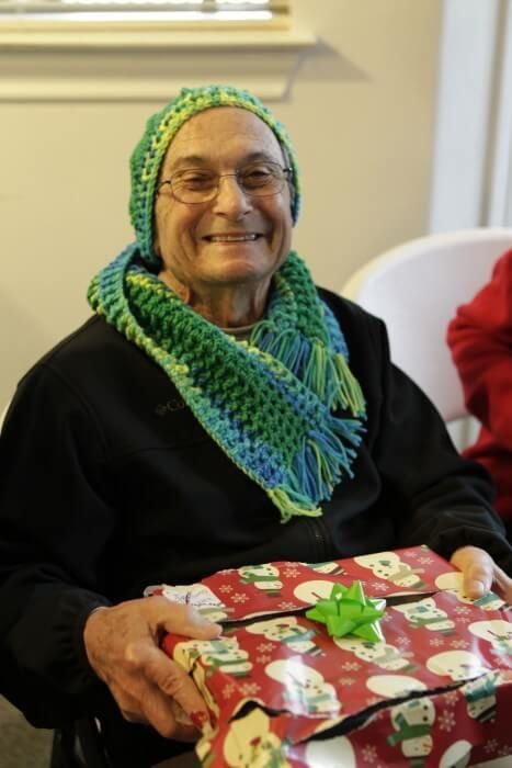 Person wearing a green and blue crocheted hat and scarf, smiling, holding a wrapped gift with a green bow.