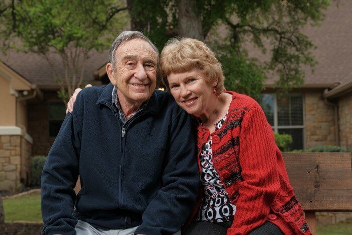 Smiling couple seated outdoors, man with arm around woman; house and tree in background.