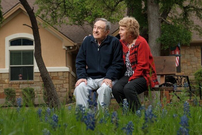 Couple sitting on a bench in front of a house, surrounded by blue flowers.