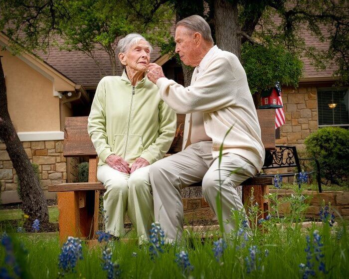 Elderly couple outdoors, man touches woman's face while sitting on a bench; house and flowers in background.