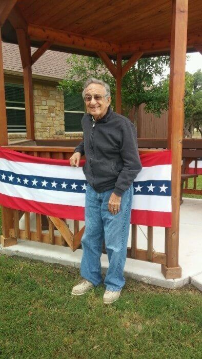 Man in a dark jacket and jeans, standing near an American flag banner on a wooden structure.