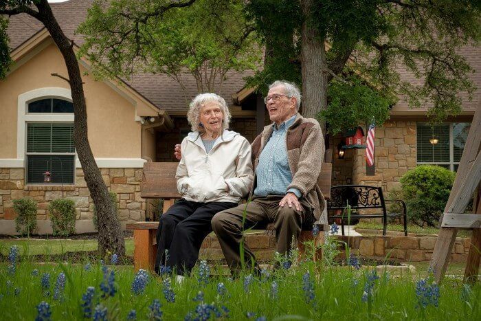 Elderly couple sitting on a bench, one with arm around the other, in a yard with a house in the background.