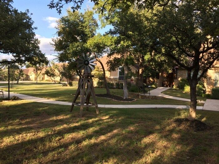 Grassy yard with trees, a small windmill, and a walkway leading to a building. Sunlight casts shadows.