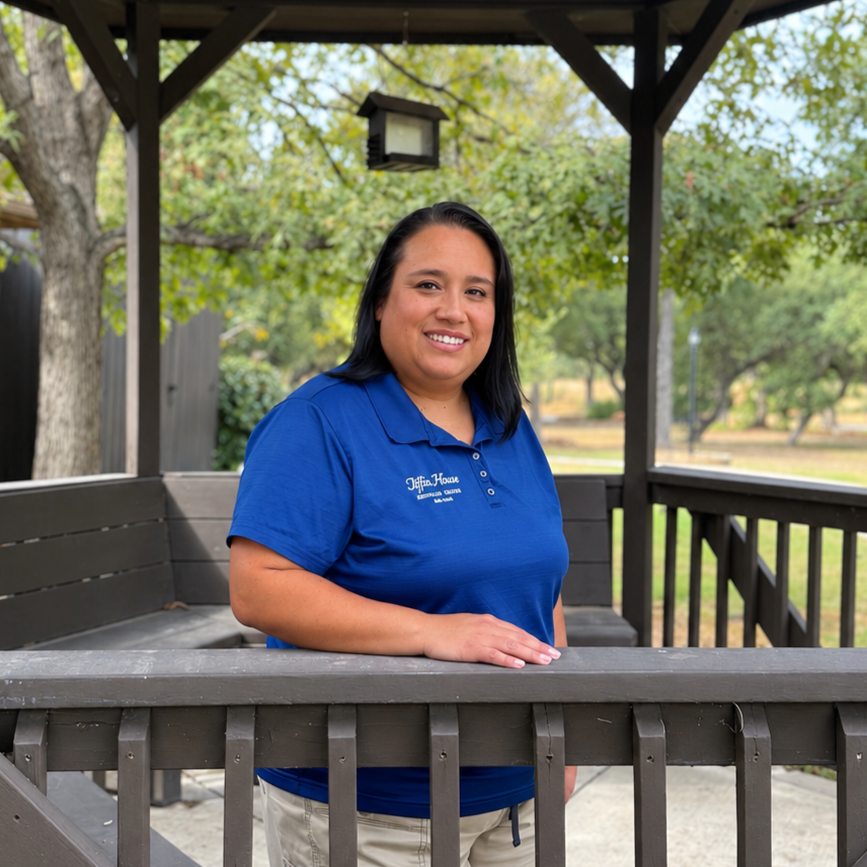 Smiling woman in a blue polo leaning on a porch railing in a park-like setting