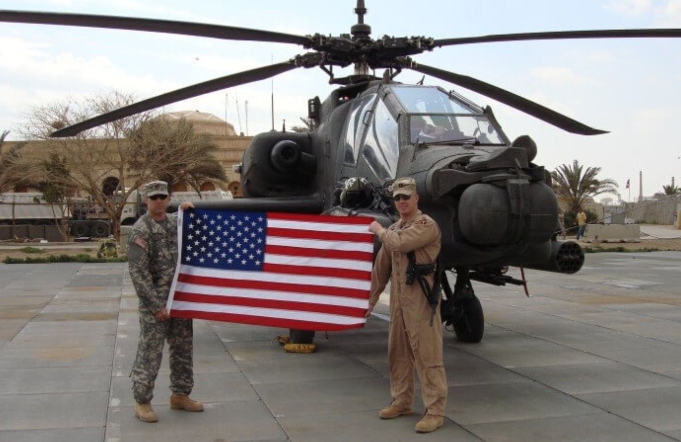 Two soldiers holding an American flag in front of an Apache helicopter.