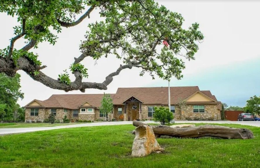 Large ranch-style building with a brown roof and stone facade, under a large tree with a green lawn.