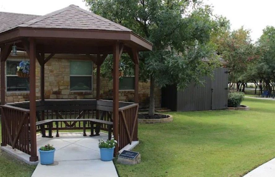 Brown gazebo with built-in benches, blue potted flowers, and a grassy yard.