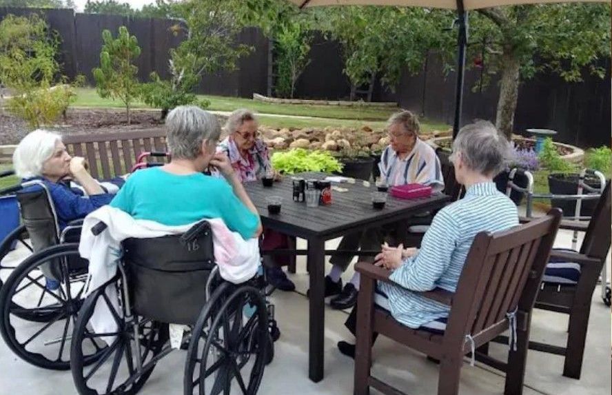 Group of people seated at an outdoor table; one person in a wheelchair. They are socializing, with a patio and garden in the background.