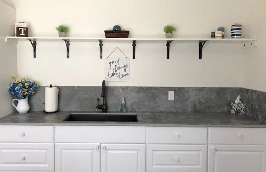 White cabinetry with a gray countertop and backsplash, black sink and faucet. Floating shelf holds decor.