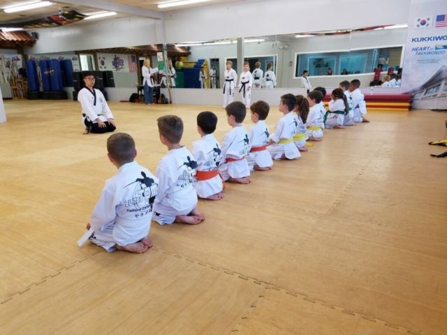 Children in martial arts uniforms kneeling, facing instructor in dojo.