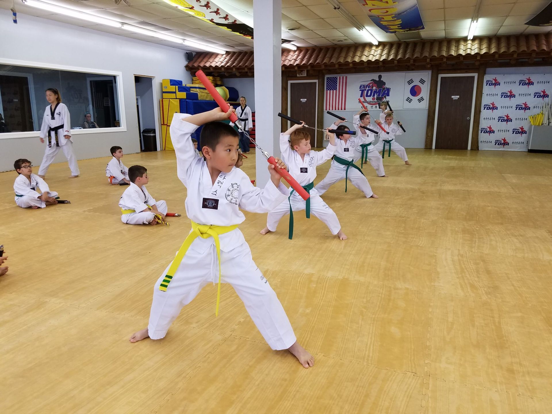 Boy in yellow belt, holding nunchucks, practices martial arts. Other children, white uniforms, training in a dojo.