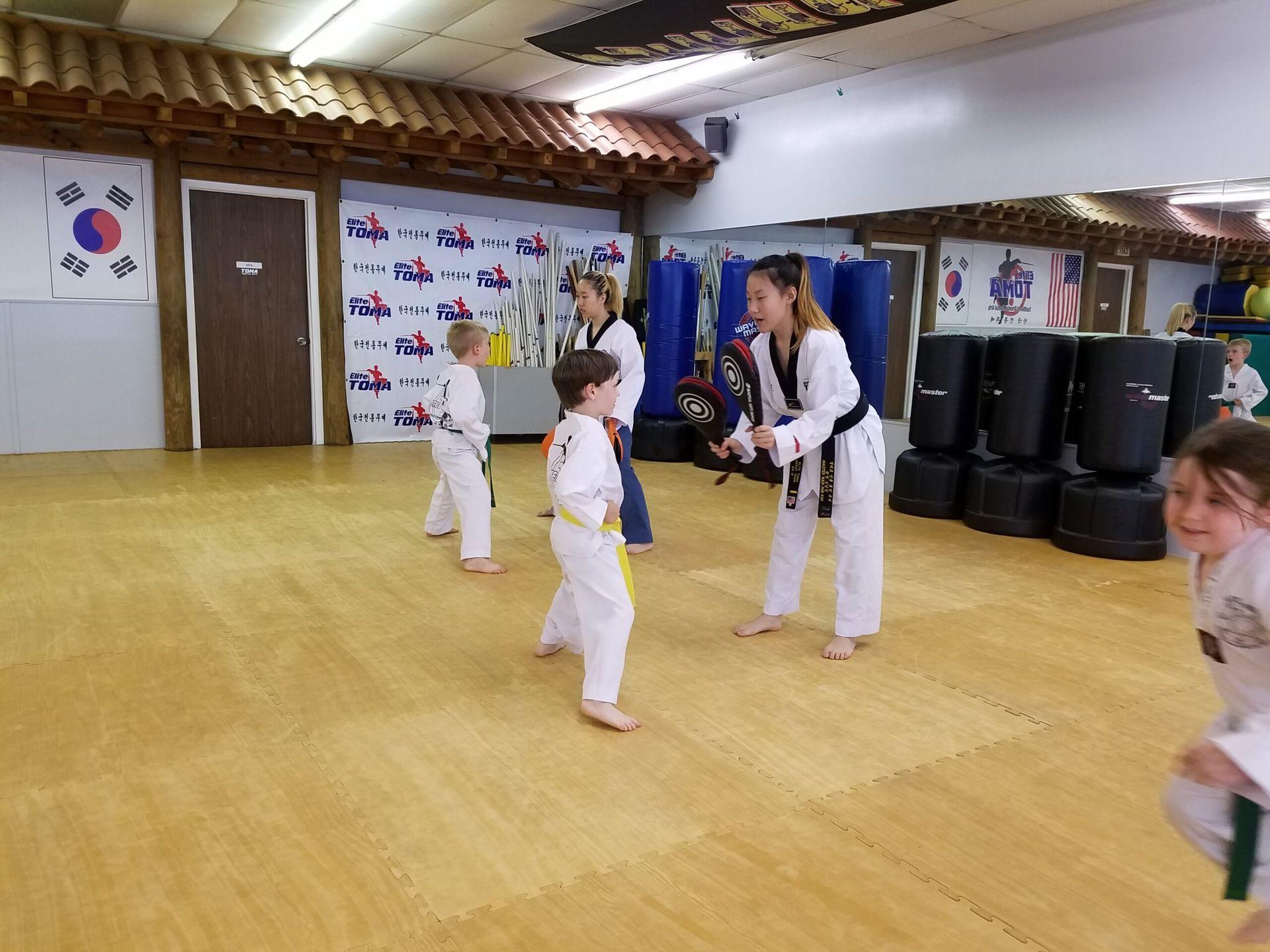 Taekwondo class in progress. Students in white uniforms practice with instructors on a yellow mat.