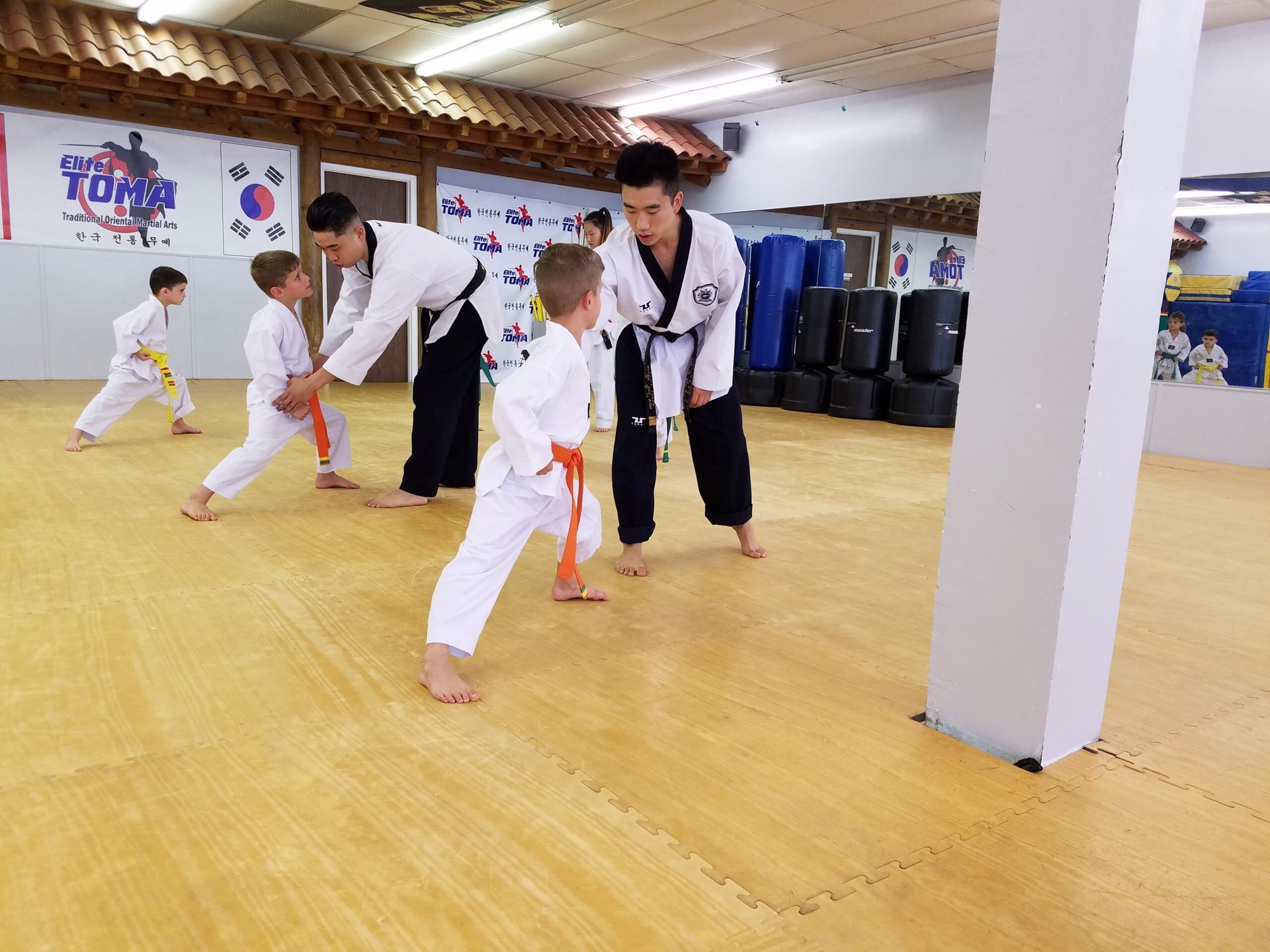 Children in white karate uniforms practice with instructors in a dojo with wooden trim.
