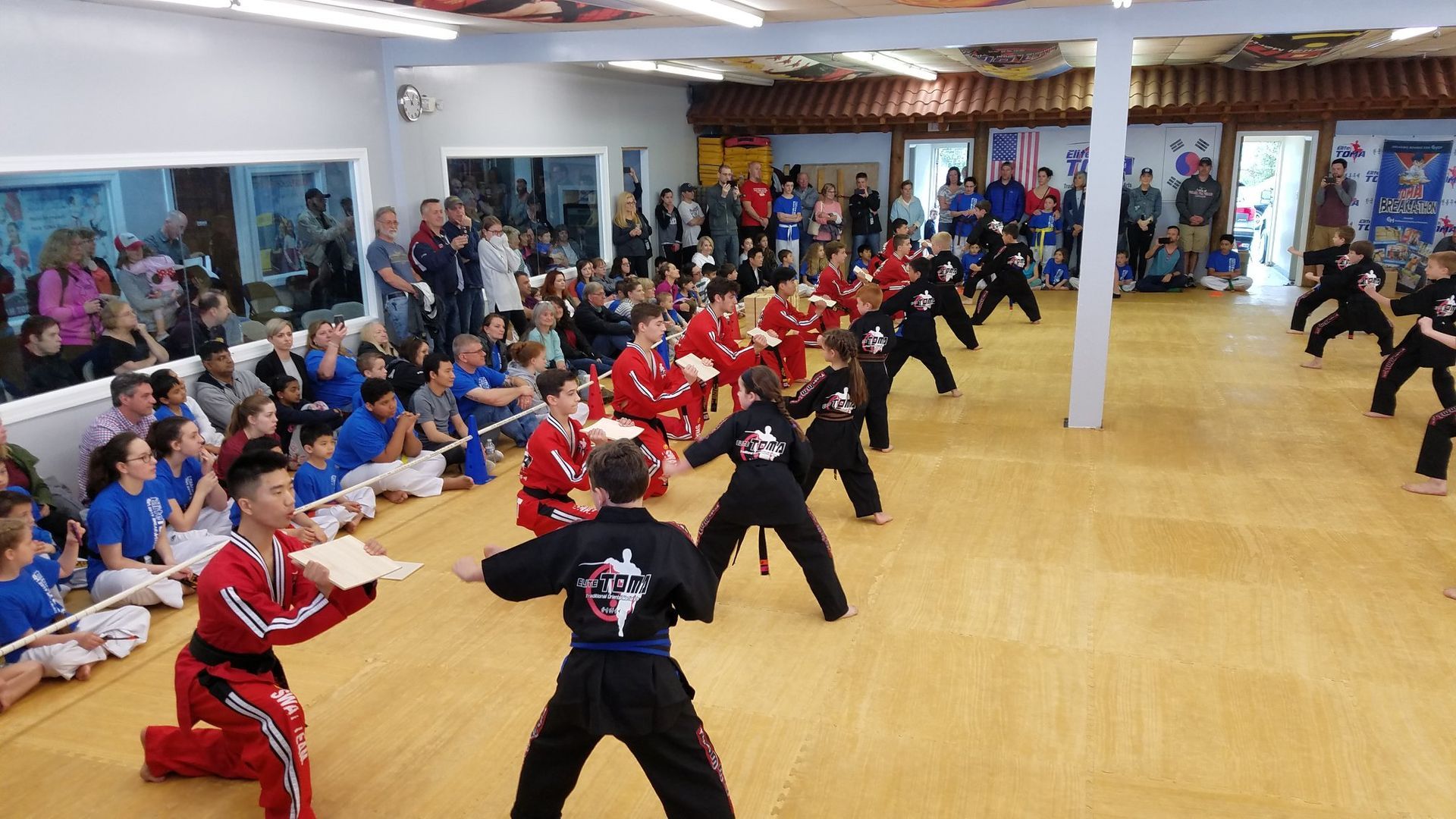 Martial arts students performing a demonstration in a dojo. People watch in the background. Students wear black and red uniforms.