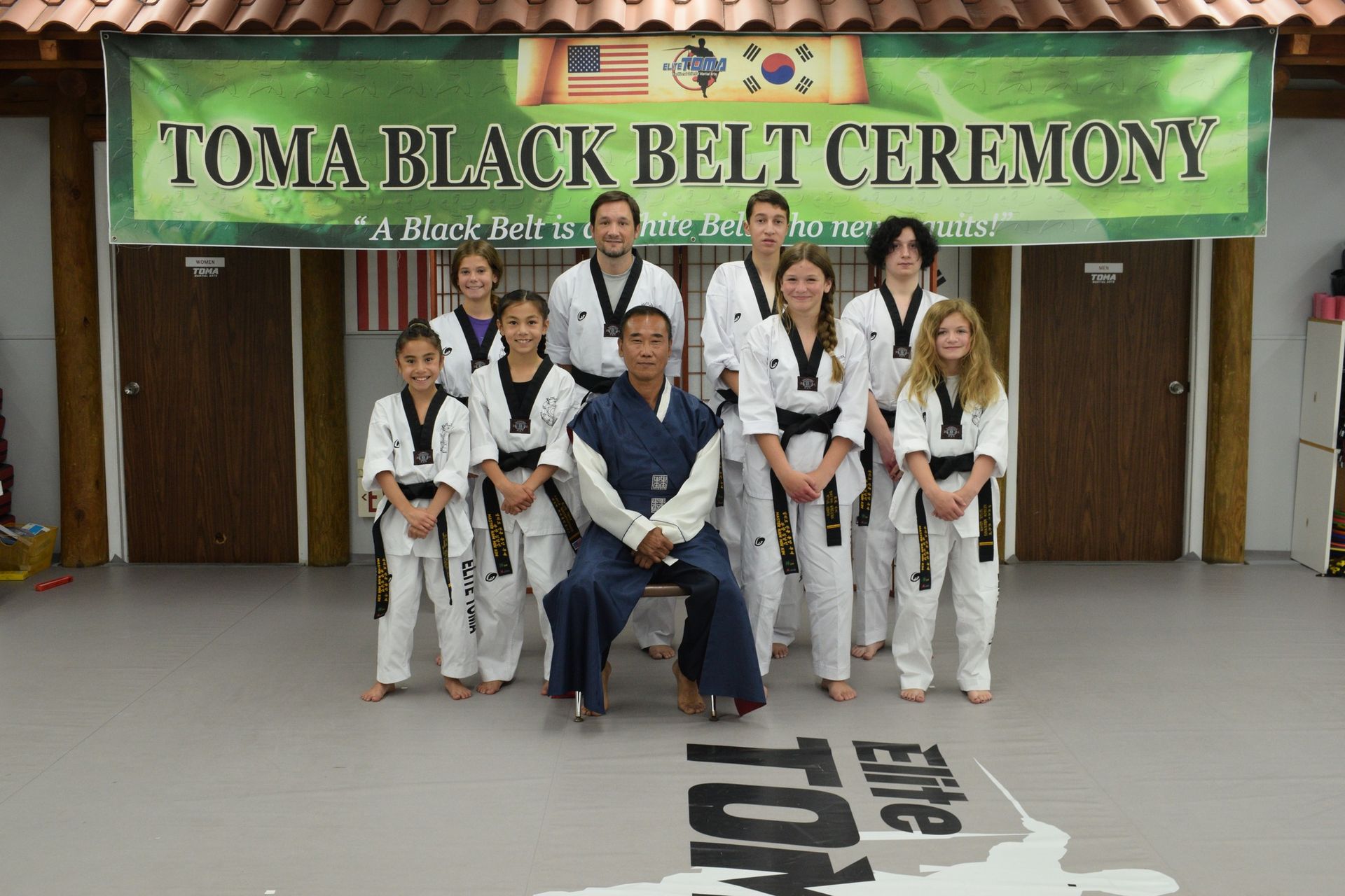 Group of people in martial arts uniforms posing for a black belt ceremony. Green banner reads 