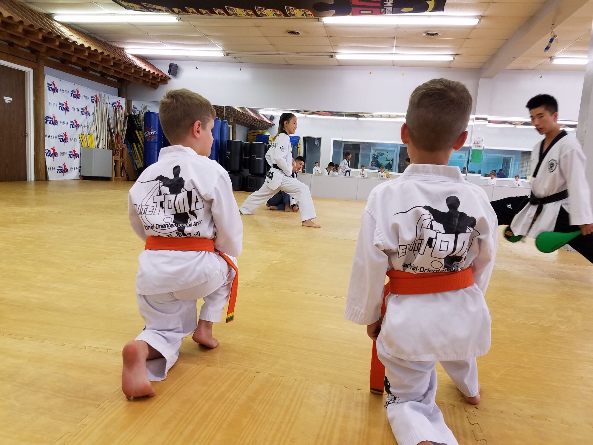 Two children in orange belts kneel on a gym floor, facing away. Others practice taekwondo in the background.