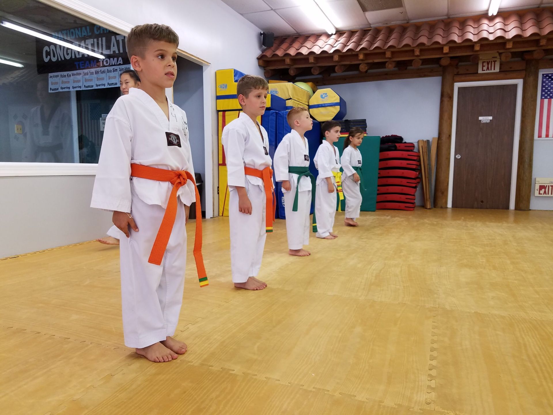 Children in martial arts uniforms with colored belts, standing in a line on a mat inside a dojo.