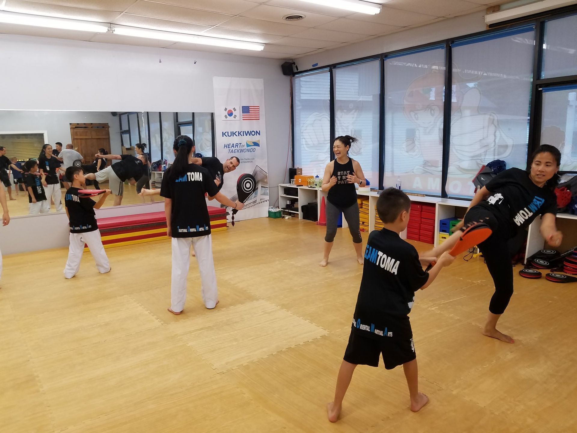 Martial arts class: Students practicing kicks, instructor holding pad. Inside a gym with mats and mirror.