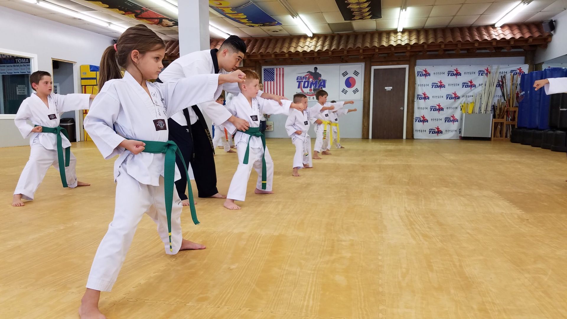 Children in white martial arts uniforms practice kicks under the instruction of a man in black attire on a wood floor.
