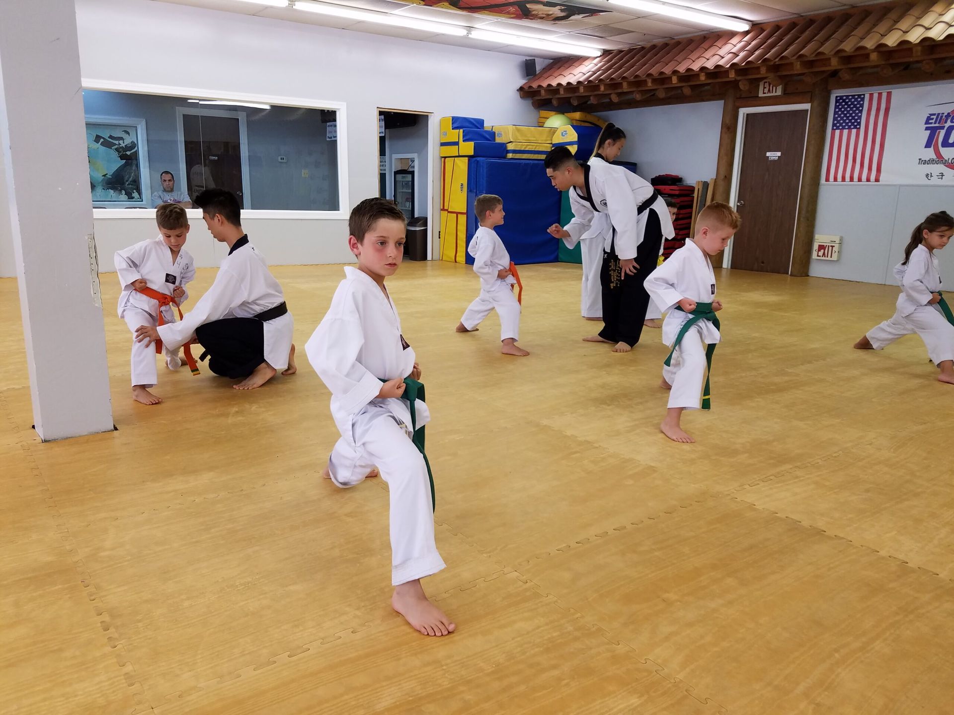 Children in white martial arts uniforms practice kicks and stances on a yellow mat. A man instructs.