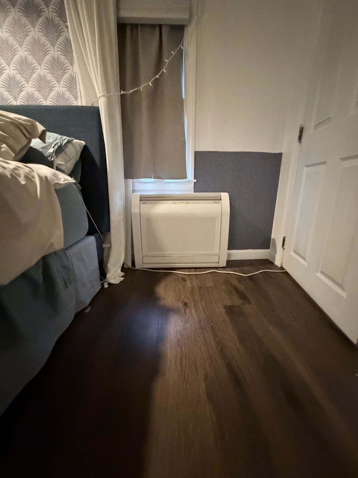 Small bedroom with bed, window and curtain, white door, and portable AC unit on the floor.
