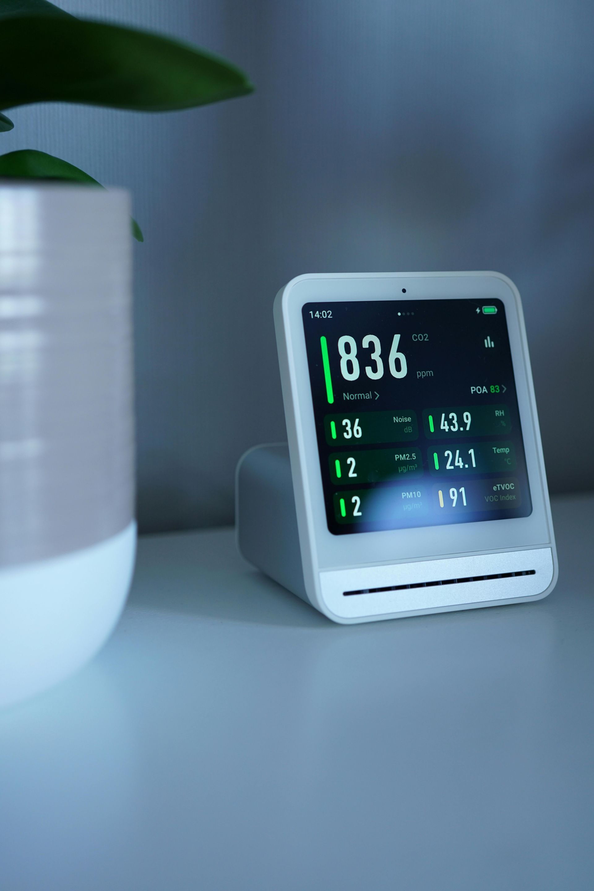Digital bedside clock on a gray table next to a white plant pot, showing 8:36 AM and weather data.