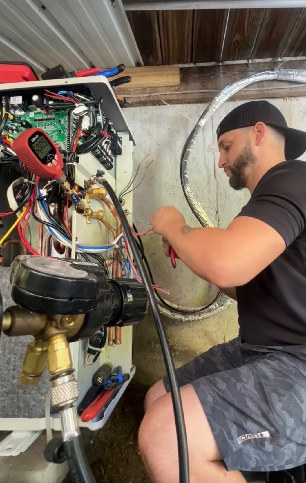 Technician working on exposed plumbing and wiring beside a large mechanical unit in a basement.