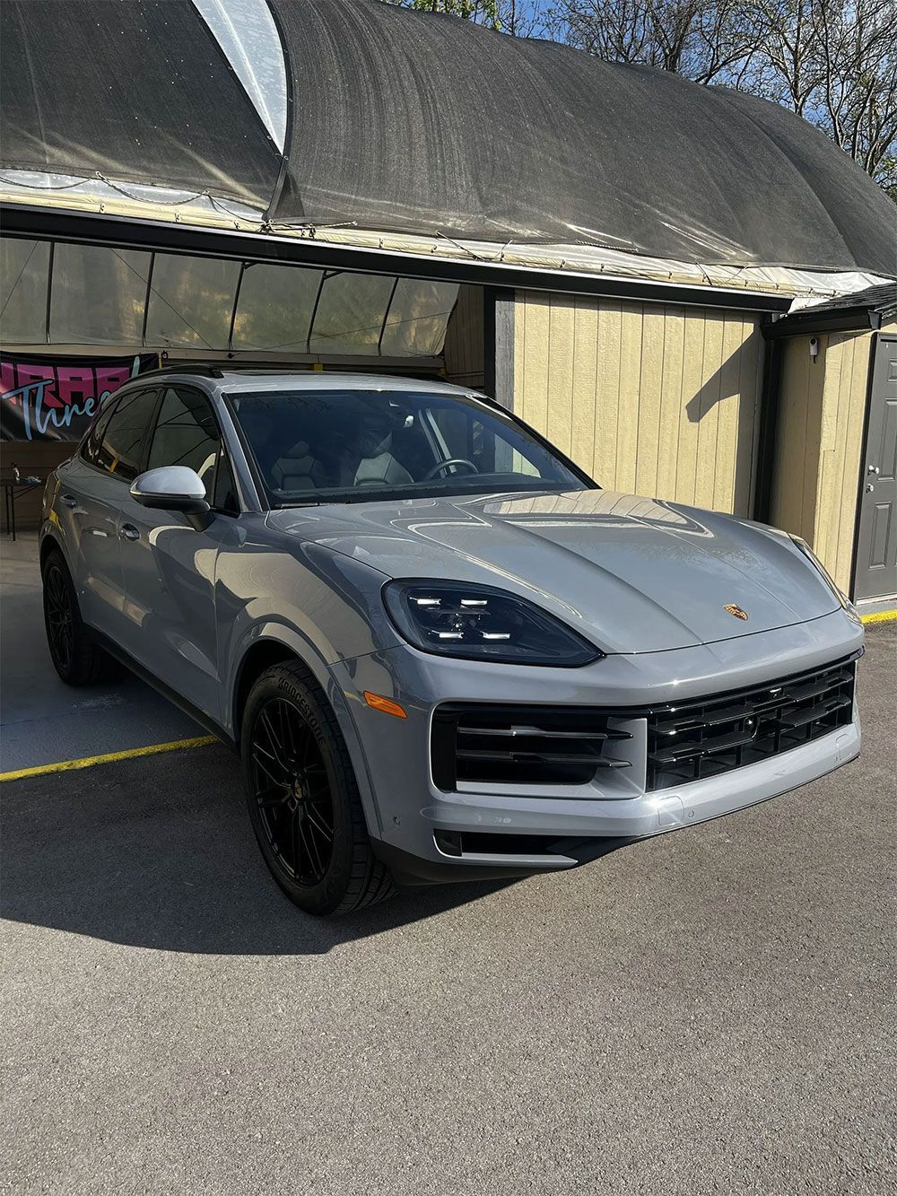 A gray Porsche Cayenne SUV parked outdoors in front of a building with a curved, shaded entrance.