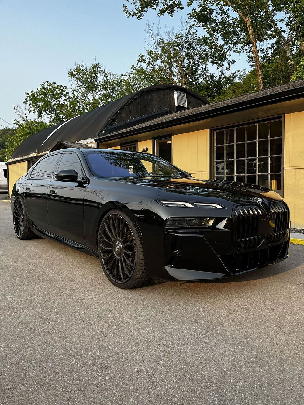 A sleek, glossy black BMW luxury sedan parked outdoors on a paved surface in front of a tan building with trees.