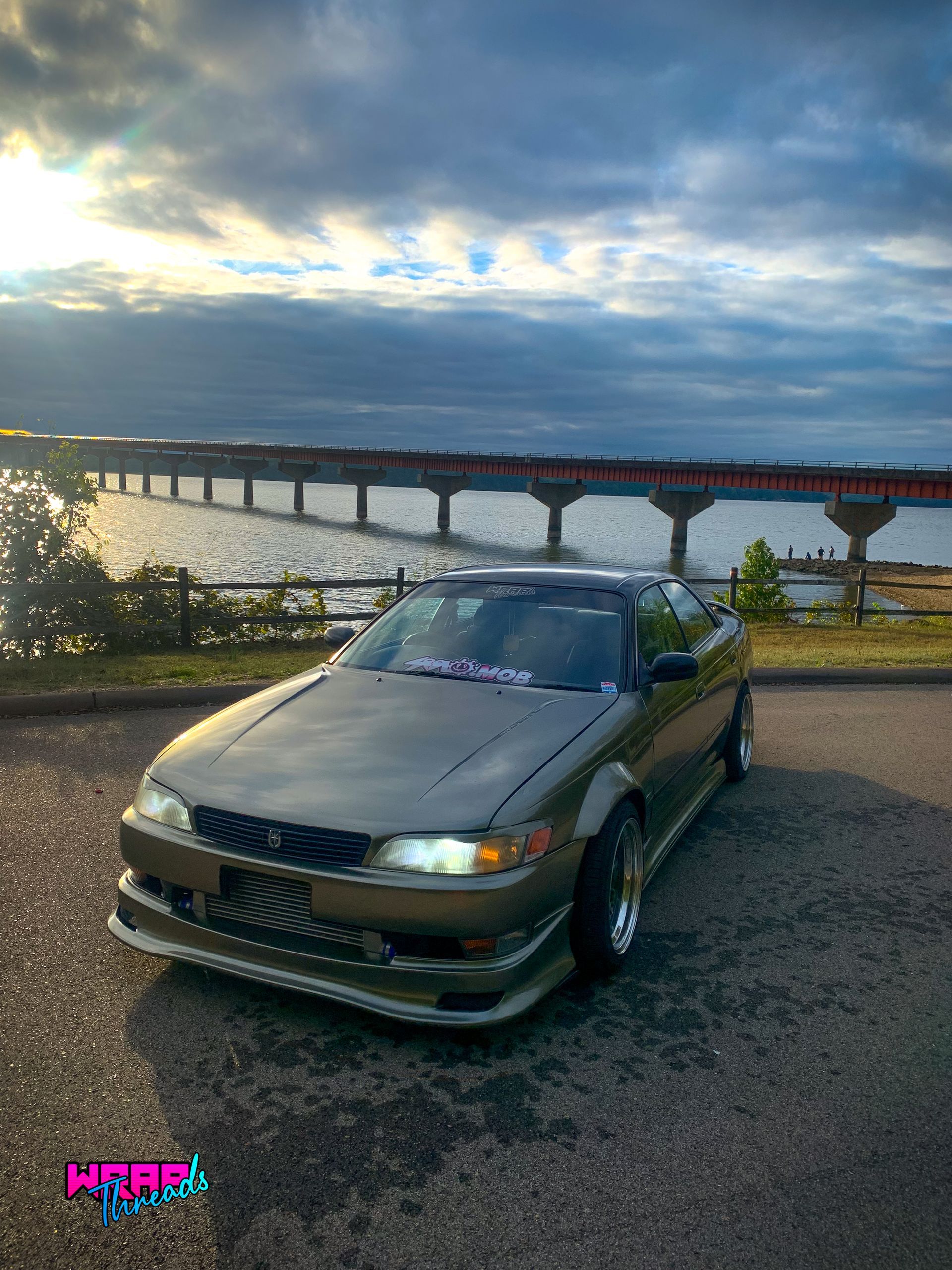A bronze Toyota Mark II parked near a bridge over water at sunset, with a prominent front intercooler visible.
