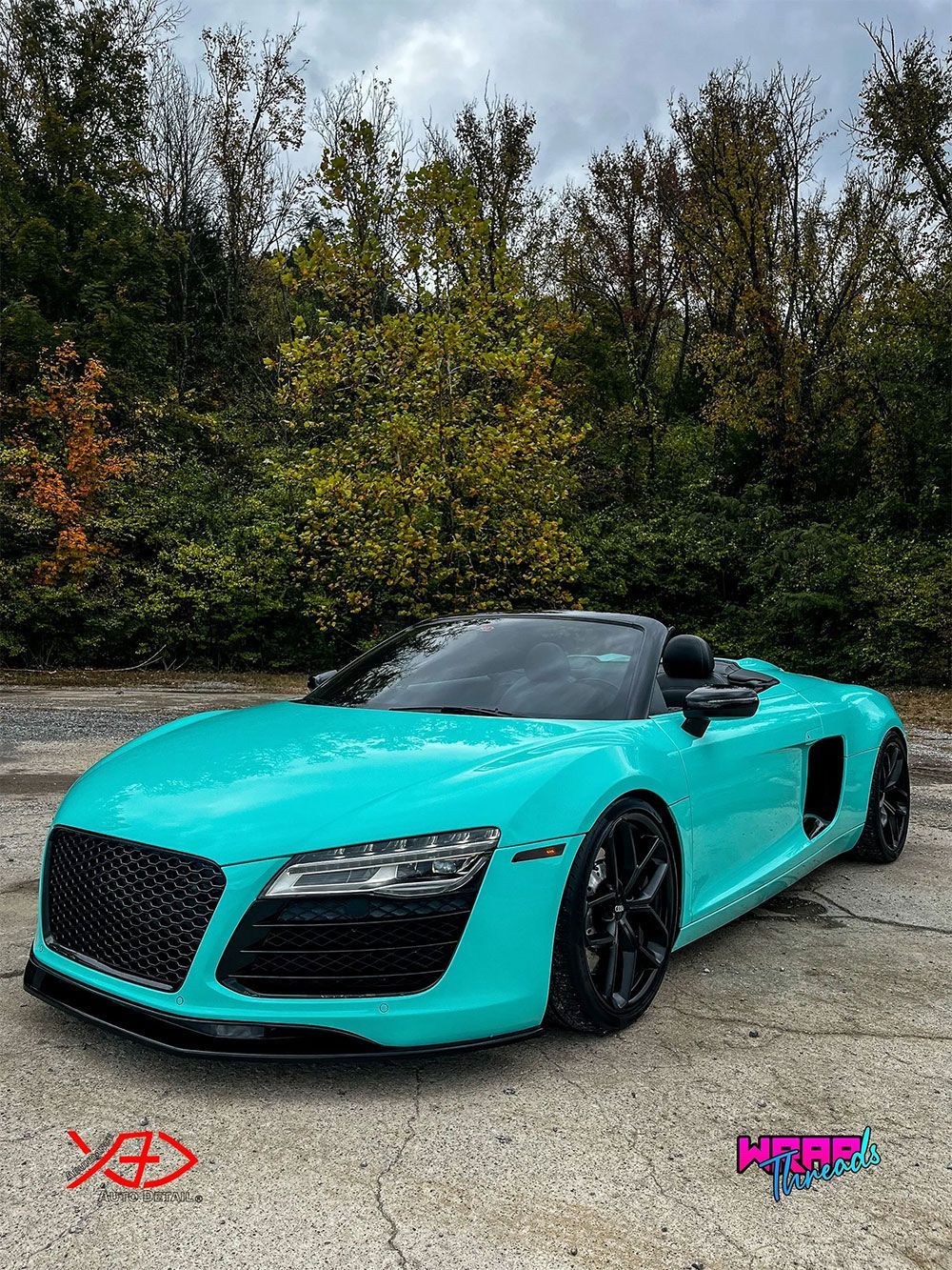A bright teal Audi R8 convertible parked on a paved surface in front of a line of trees.