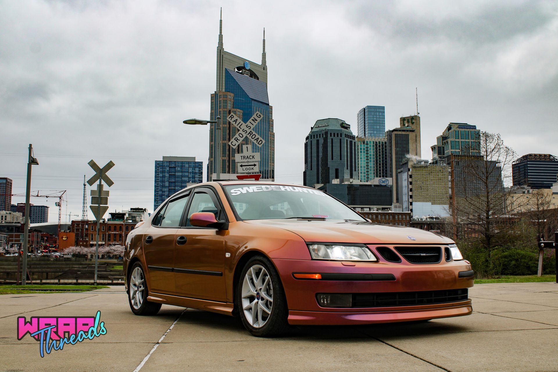 A color-shifting orange and purple Saab parked in front of the Nashville skyline with a railroad crossing sign.