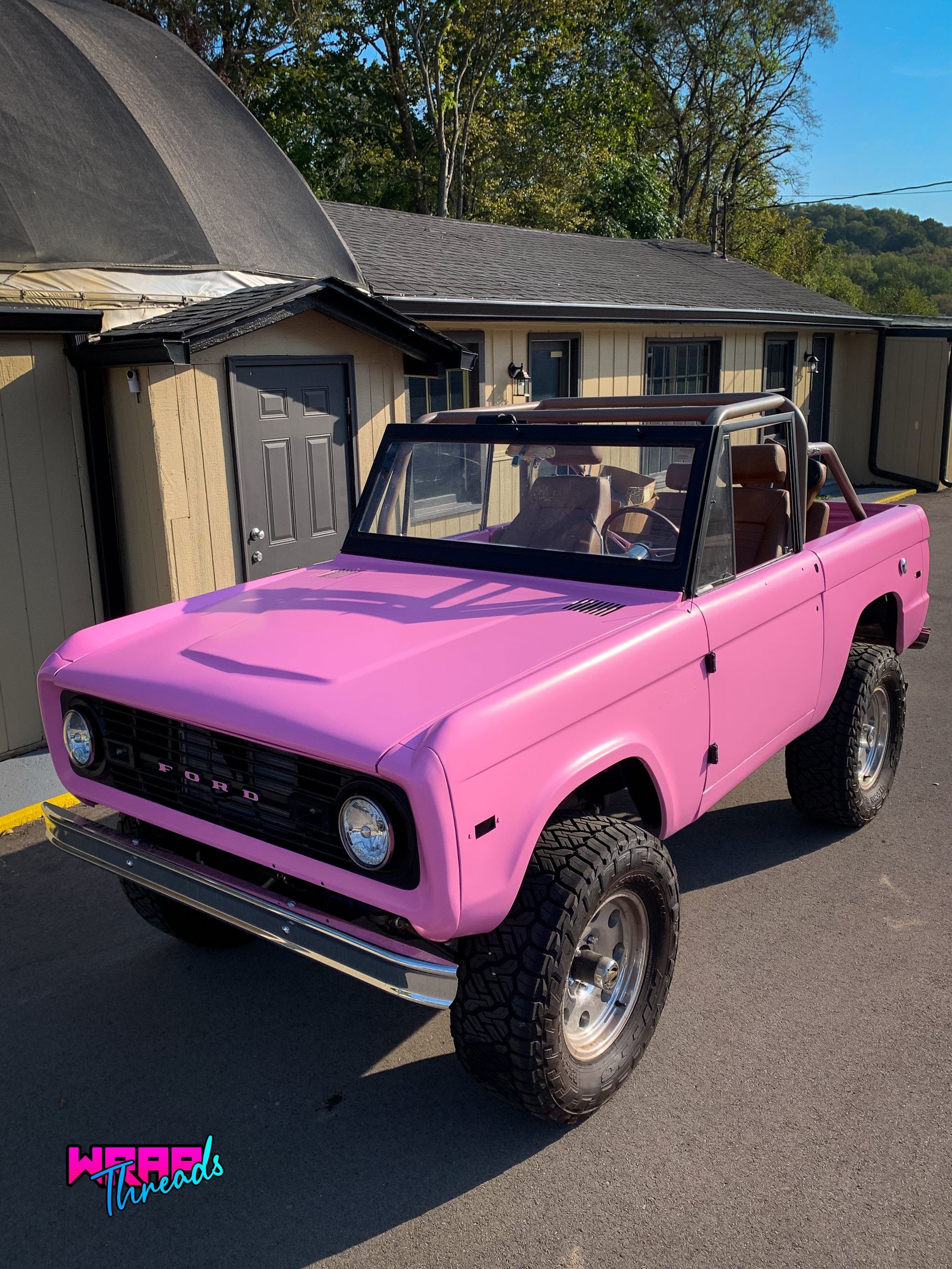 A bright pink vintage Ford Bronco parked outdoors in front of a building on a sunny day.