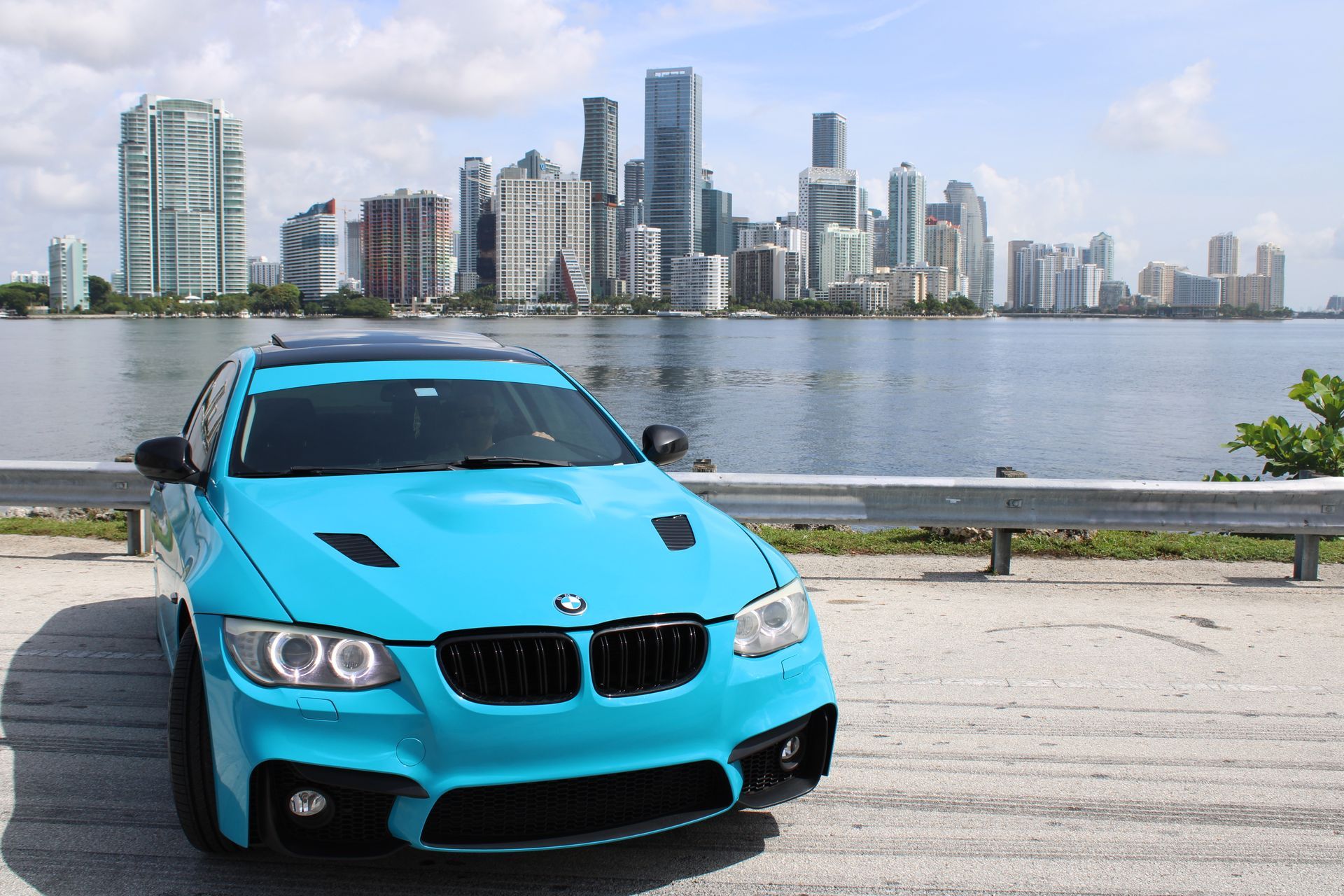 A bright blue BMW sports car parked along a waterfront with a city skyline in the background.