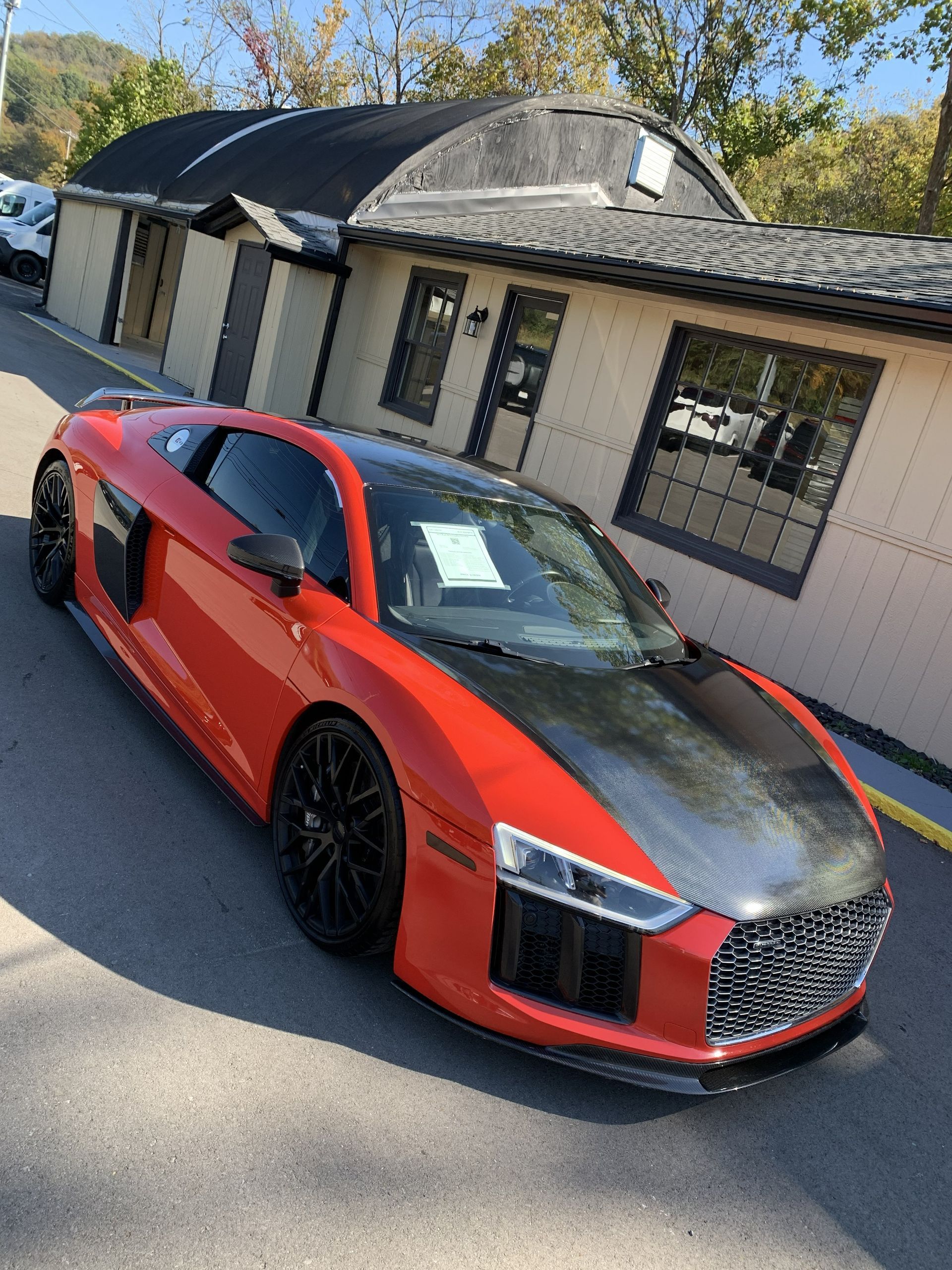 A bright orange Audi R8 with a black hood and black wheels parked in front of a tan, single-story commercial building.