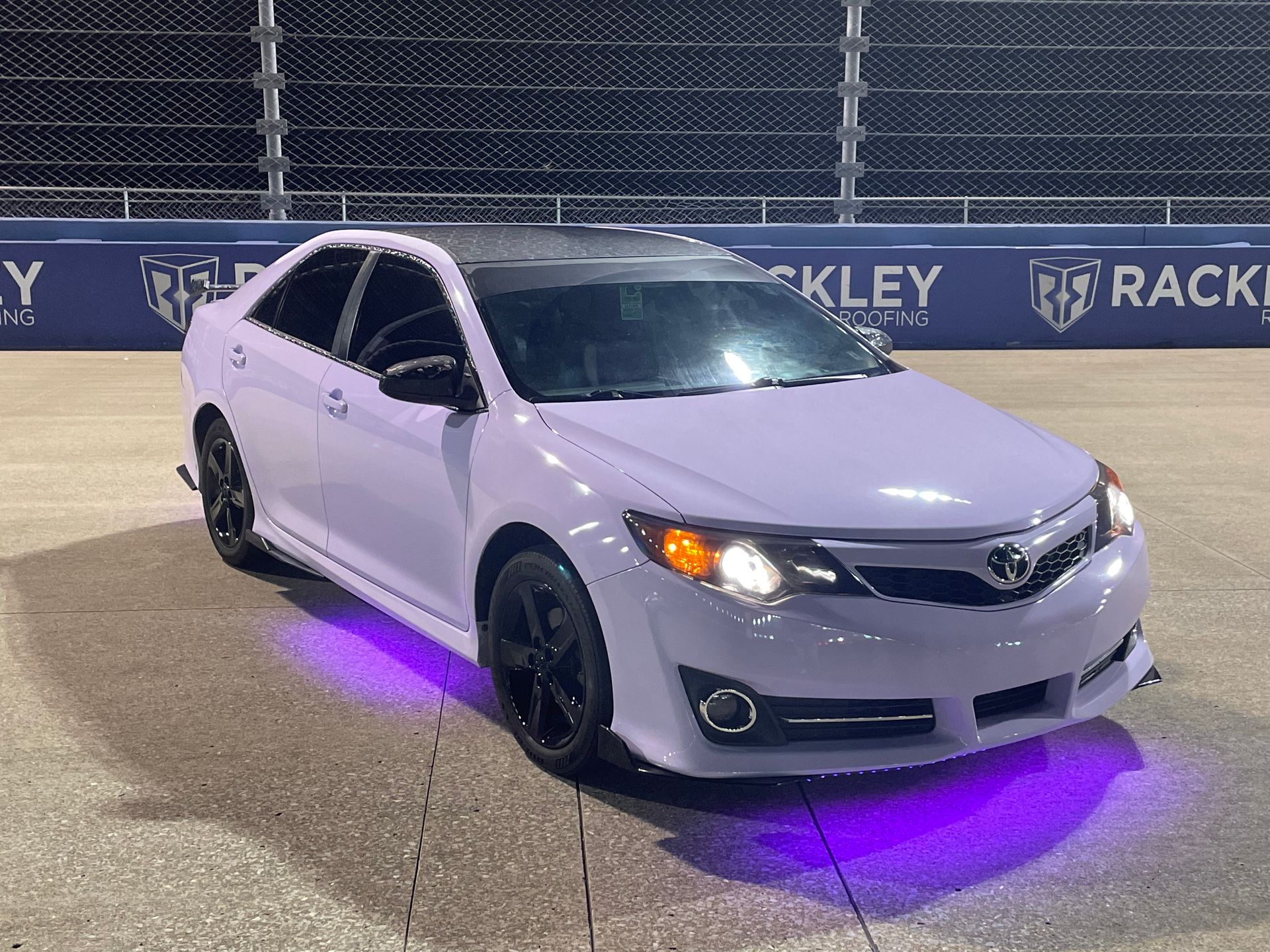 A lavender sedan with black wheels and purple underglow parked on a track at night, with Rackley signage in the background.