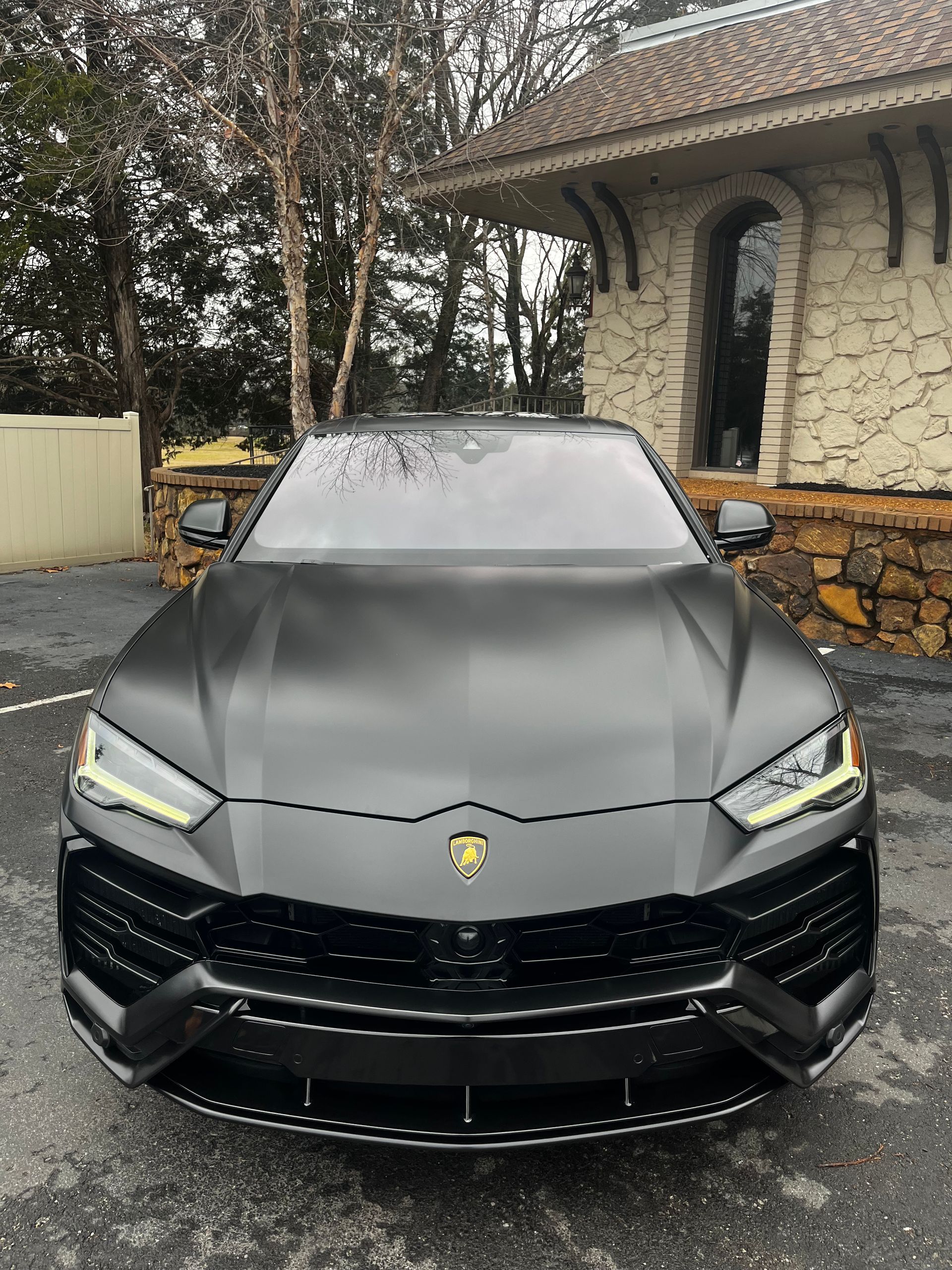 A matte black Lamborghini Urus SUV parked in front of a stone building with trees in the background.