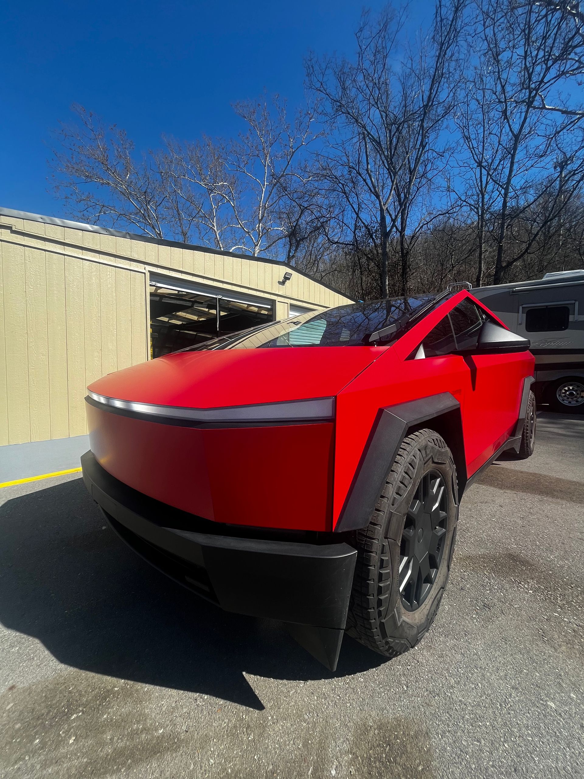 A vibrant red Tesla Cybertruck with black wheel arches and wheels parked on a gravel lot under a clear blue sky.
