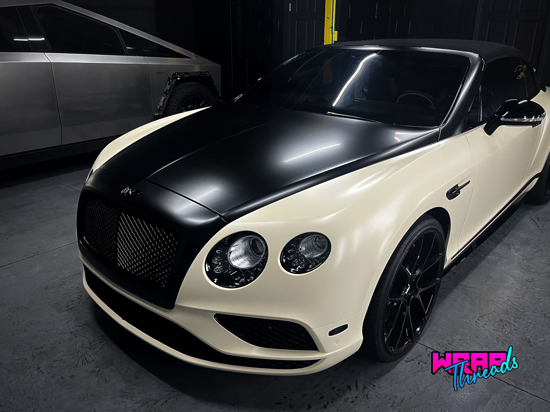 A two-tone Bentley with a matte black hood and grille and cream-colored body, parked next to a silver vehicle.