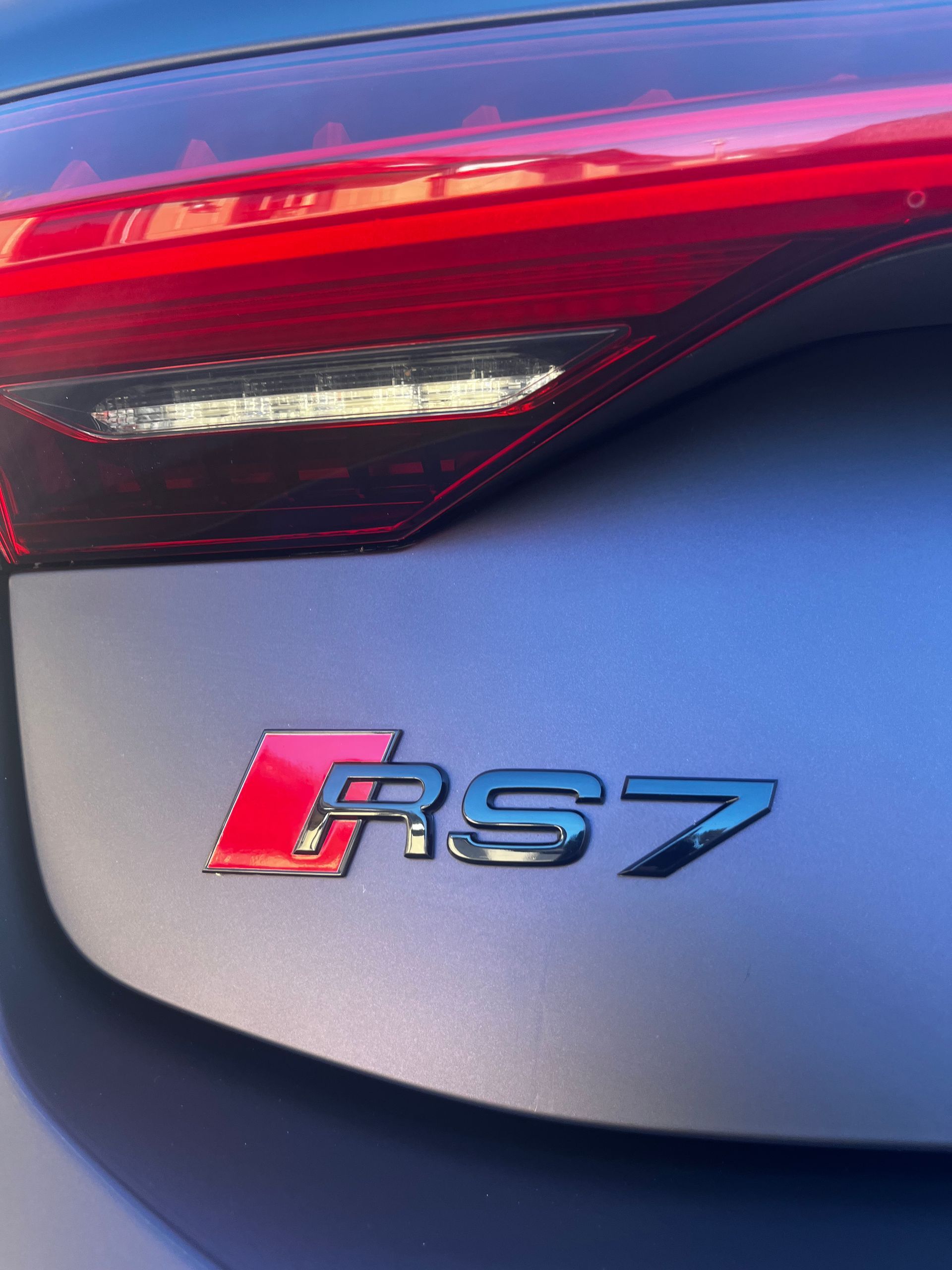 Close-up of a black Audi RS7 badge on a matte gray car trunk, positioned below the red LED taillight.