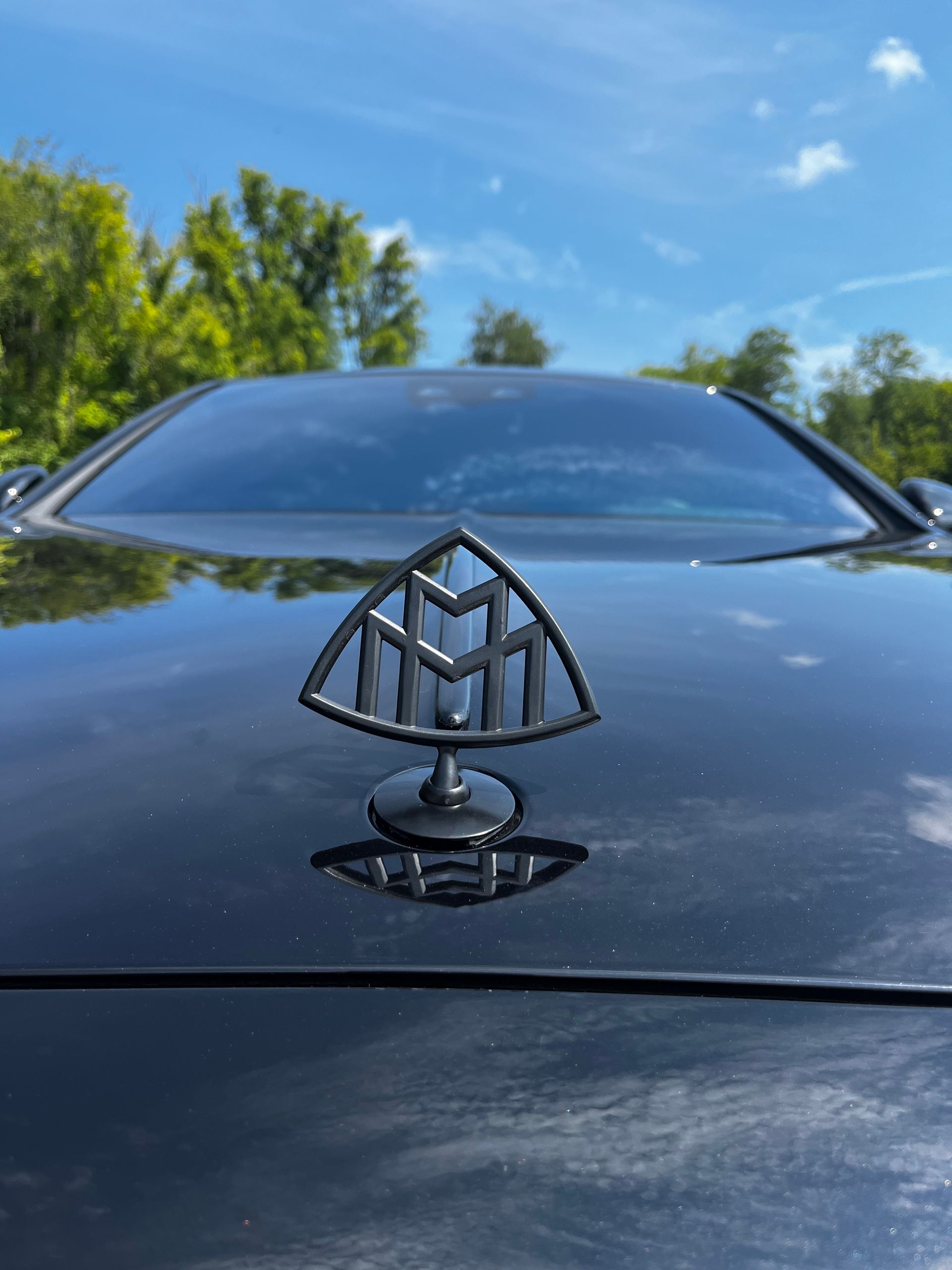 A black Maybach hood ornament mounted on a shiny black car hood against a blue sky with green trees.